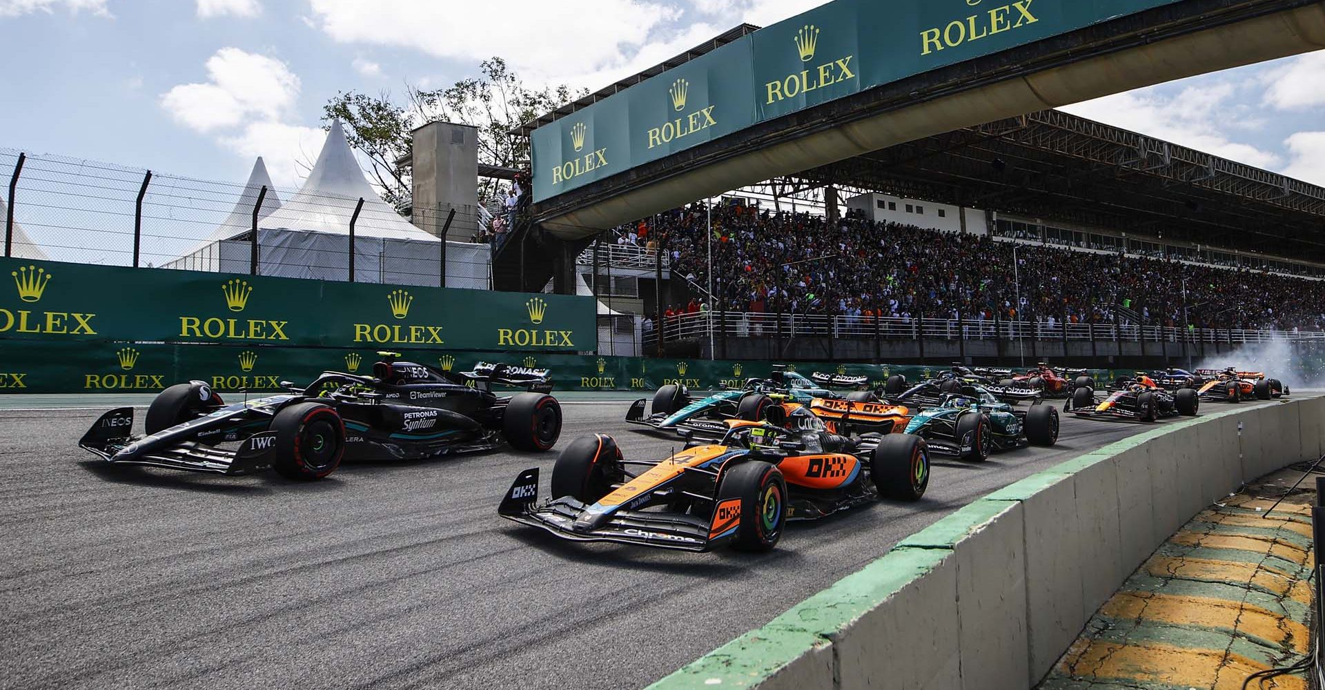 Lando Norris, McLaren MCL60, leads Sir Lewis Hamilton, Mercedes F1 W14, Fernando Alonso, Aston Martin AMR23, and Lance Stroll, Aston Martin AMR23, at the start as Kevin Magnussen, Haas VF-23, and Alex Albon, Williams FW45, crash out in the background